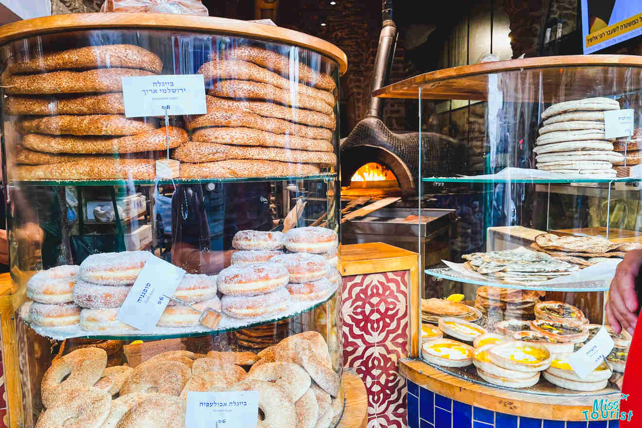 A bakery display case filled with assorted bagels and pastries, with labels in hebrew, viewed through a shop window.