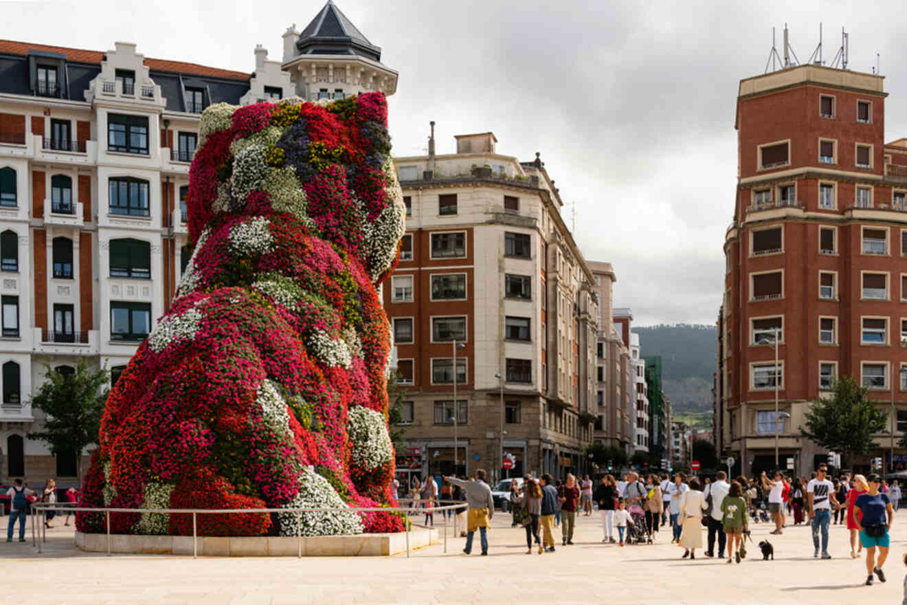 Large floral sculpture of a dog in an urban square, surrounded by pedestrians enjoying the view.