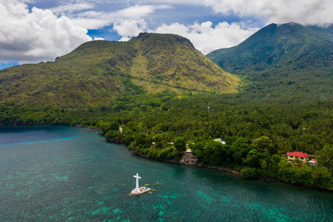 Aerial view of a coastal landscape with lush green mountains, a large cross monument in the sea, and a dense forest on the shore.