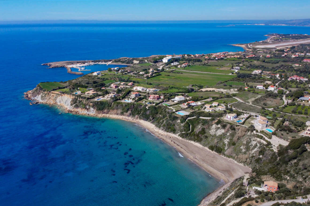Aerial view of a coastal landscape with a sandy beach, clear blue waters, and scattered buildings surrounded by greenery.