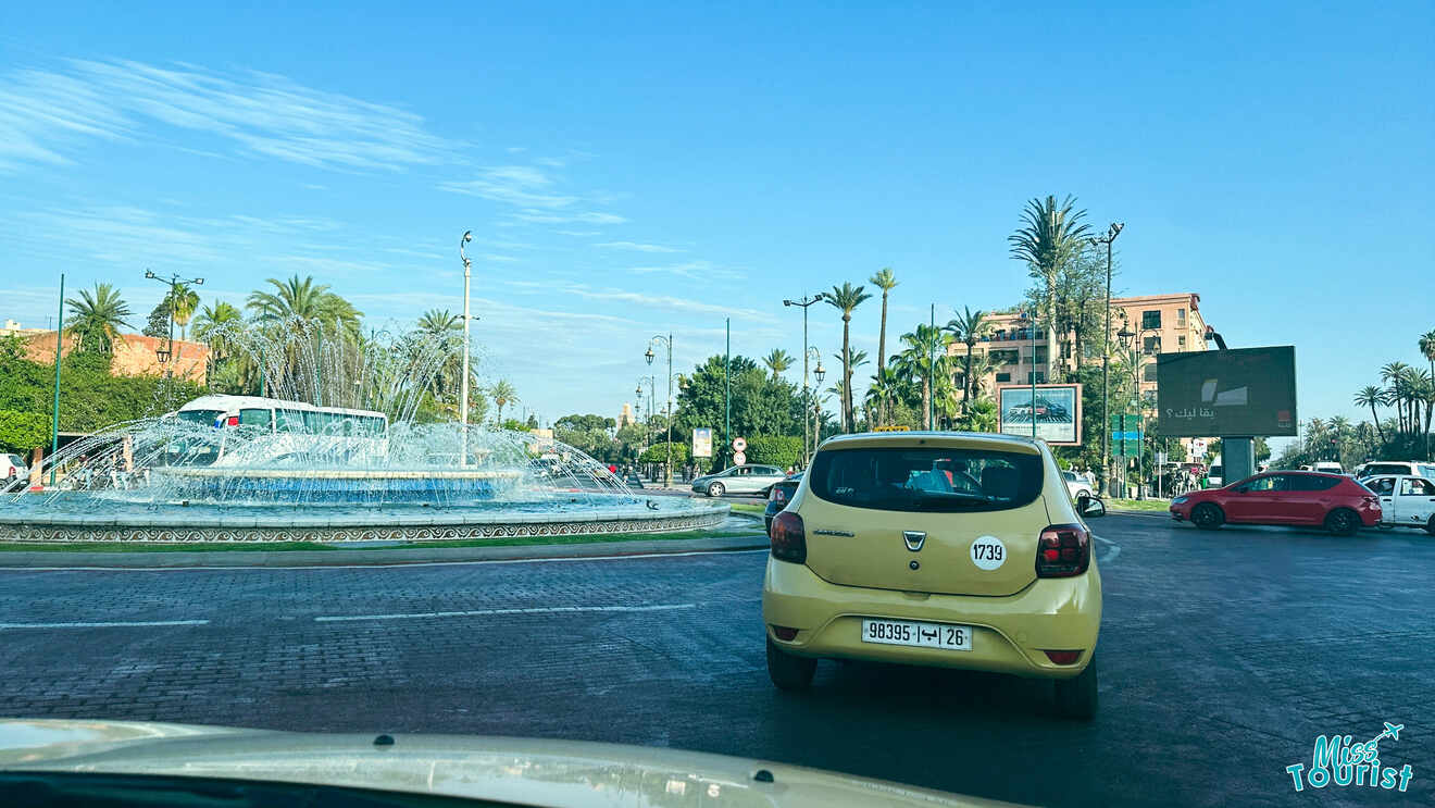 Sunny daytime traffic scene in Marrakech with cars circling a fountain at a busy intersection, showcasing the city's modern transportation amidst palm trees