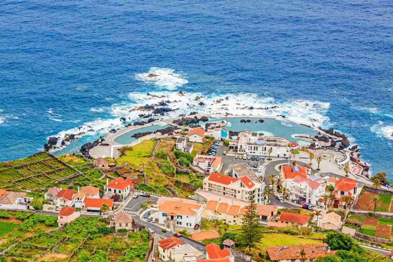 A picturesque aerial view of a coastal village in Madeira with a natural volcanic swimming pool at the seaside, surrounded by terraced fields and local architecture