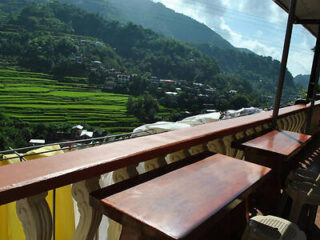 View from a balcony with wooden countertop overlooking lush green rice terraces in a mountainous area, under a clear sky.
