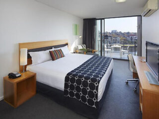 Spacious hotel room with king-sized bed with a blue comforter and balcony overlooking city skyline.