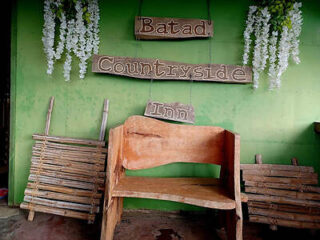 Wooden bench and chair in front of a green wall with "batad countryside inn" sign, decorated with hanging white flowers.