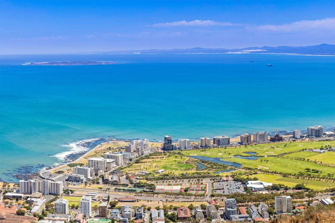 Panoramic view of a coastal Cape Town suburb with turquoise waters, green spaces, and urban development, as seen from a high vantage point