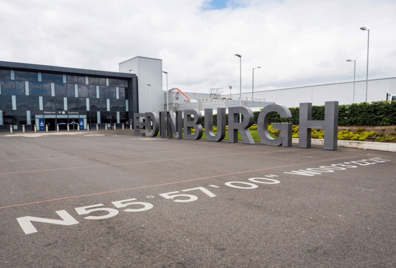 The entrance to Edinburgh Airport with a large, bold 'EDINBURGH' sign, coordinates on the tarmac, and a modern building facade, under a cloudy sky
