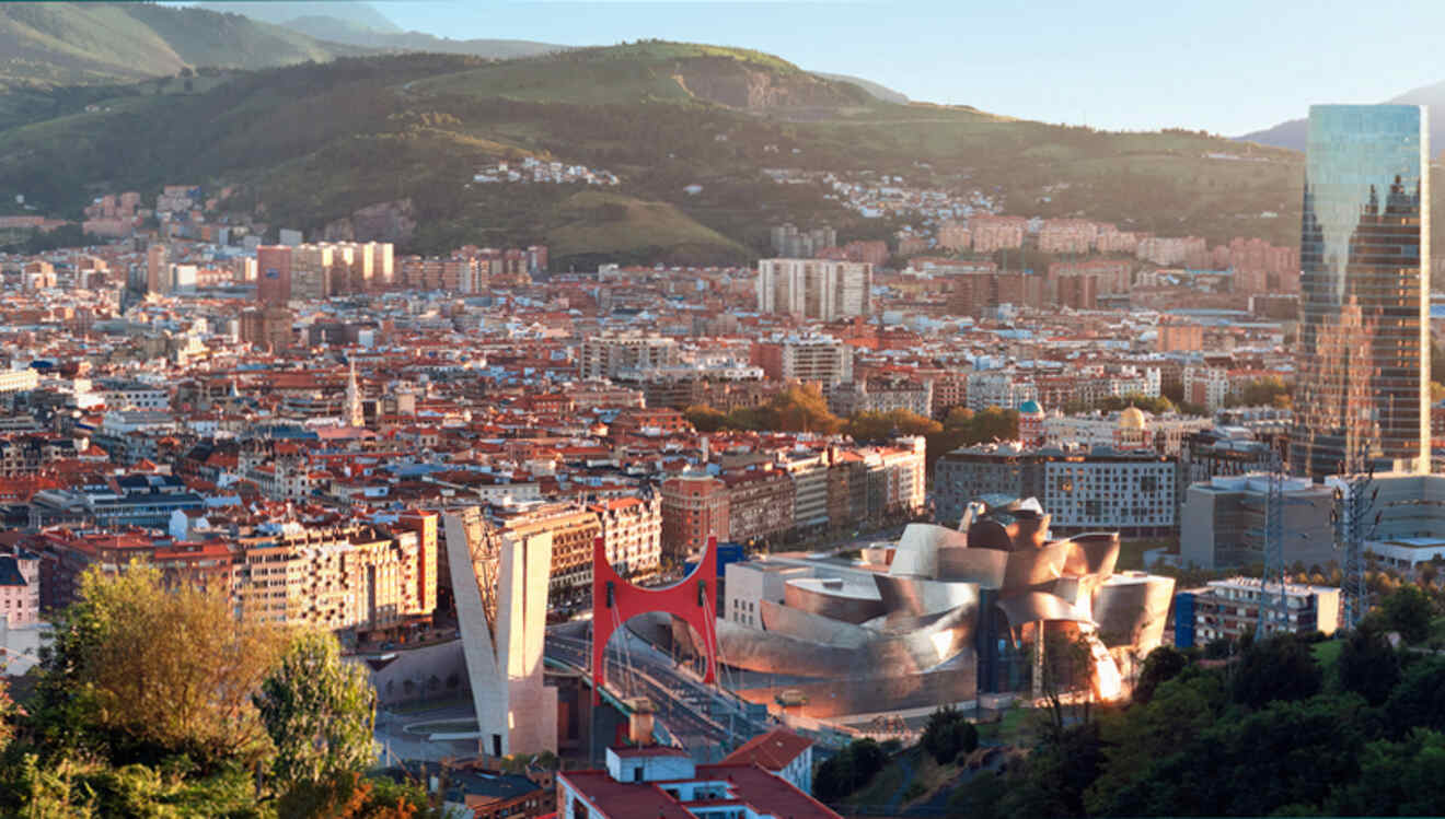 Aerial view of bilbao, spain, showcasing modern guggenheim museum and red salve bridge against a backdrop of green hills and cityscape.