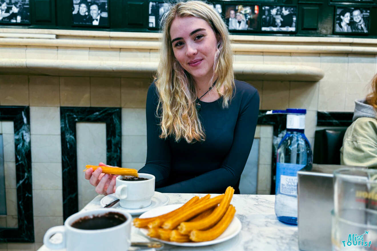 Author of the post enjoying traditional Spanish churros with chocolate at the famed Chocolatería San Ginés in Madrid, with black and white photos of celebrities adorning the tiled wall behind.