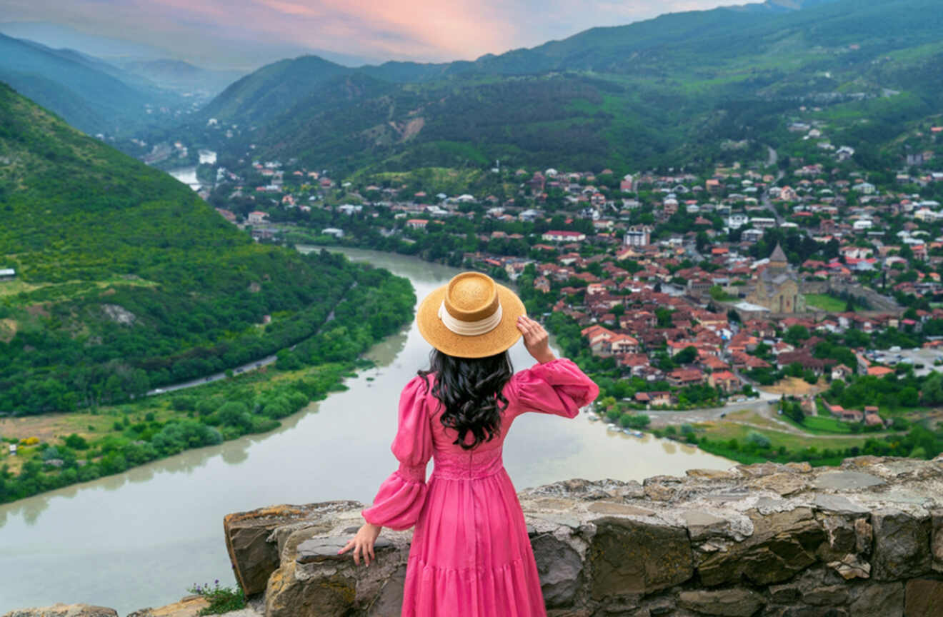 A woman in a pink dress and straw hat gazes out over the ancient city of Mtskheta, with its historical buildings and churches surrounded by the winding river and lush hills