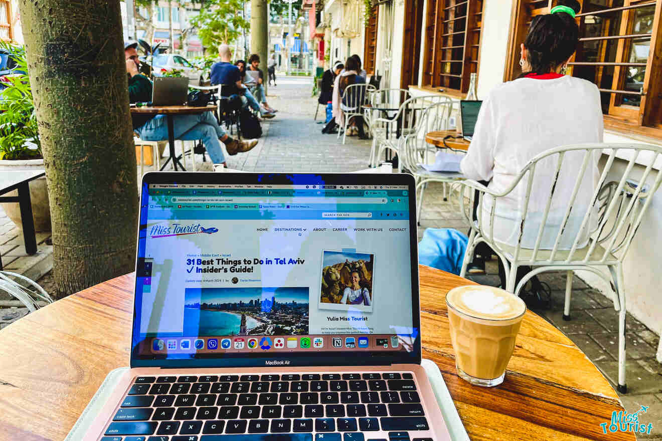 A person sitting at an outdoor café table, browsing a travel website about tel aviv on a laptop, with a cup of coffee beside the computer.
