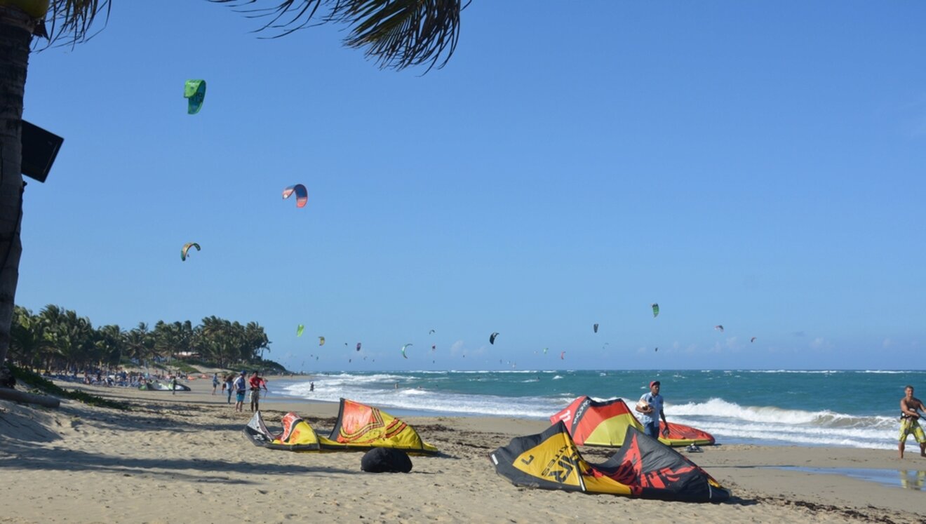 A lively beach scene in Cabarete with numerous kitesurfing parachutes in the sky and enthusiasts preparing gear on the sand.