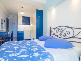 A small, efficiently designed studio apartment with a blue and white color scheme, featuring an integrated kitchen, dining area, and a bed with decorative blue bedding.