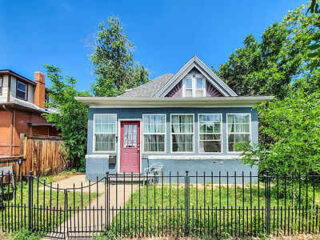 Quaint blue-gray house with white trim, red door, and a picket fence surrounded by lush greenery