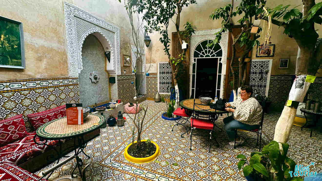 Cozy and ornate Moroccan courtyard with mosaic tiles, a seating area, and a central tree, with a guest enjoying the ambiance