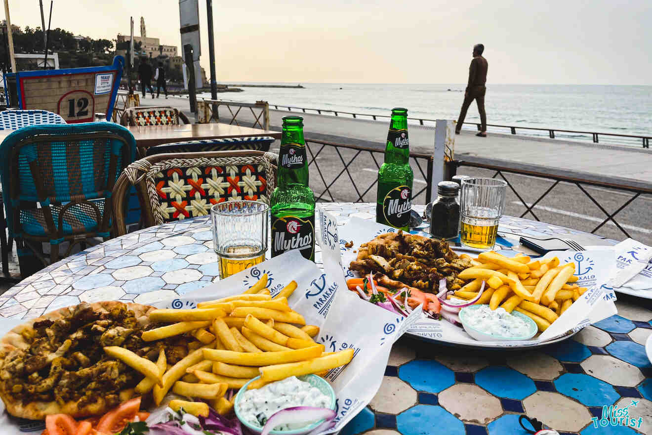 Table with seafood dishes, fries, and beers overlooking a beachside promenade with a person walking in the distance.