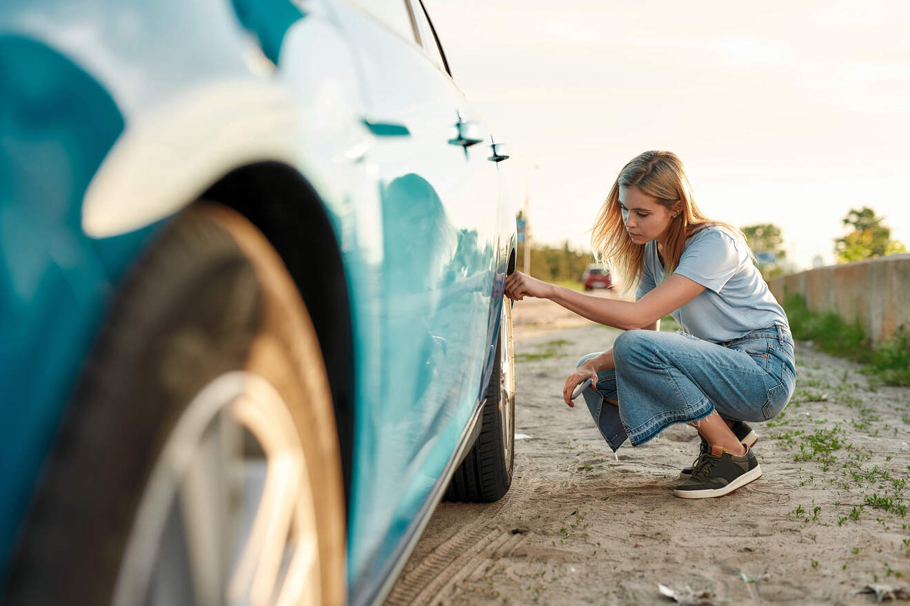 A woman crouched down checking her car tire on the side of a road, representing the importance of vehicle maintenance during travel.