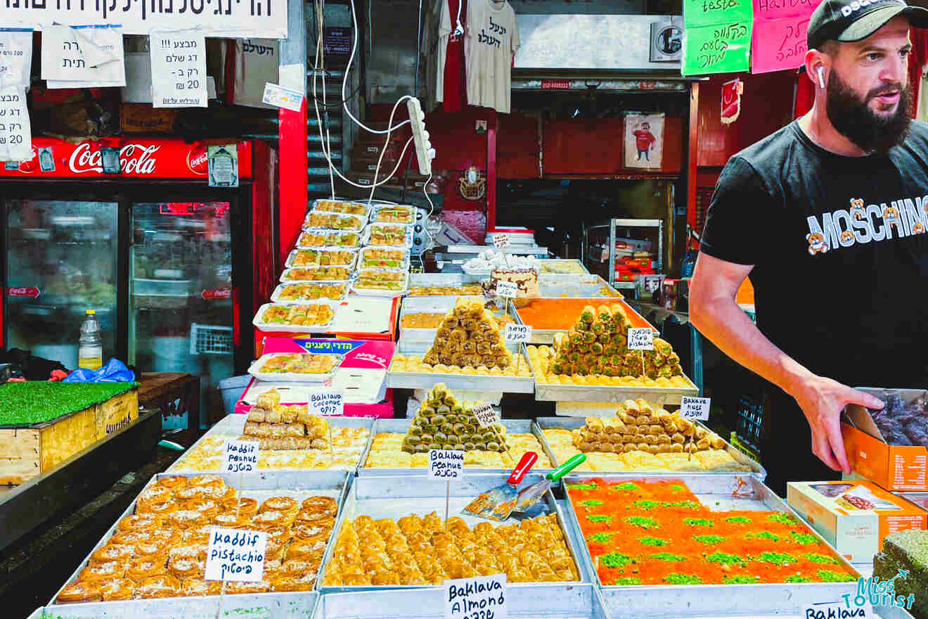 A man stands beside a variety of baklava and other sweets displayed at a vibrant market stall with price tags.