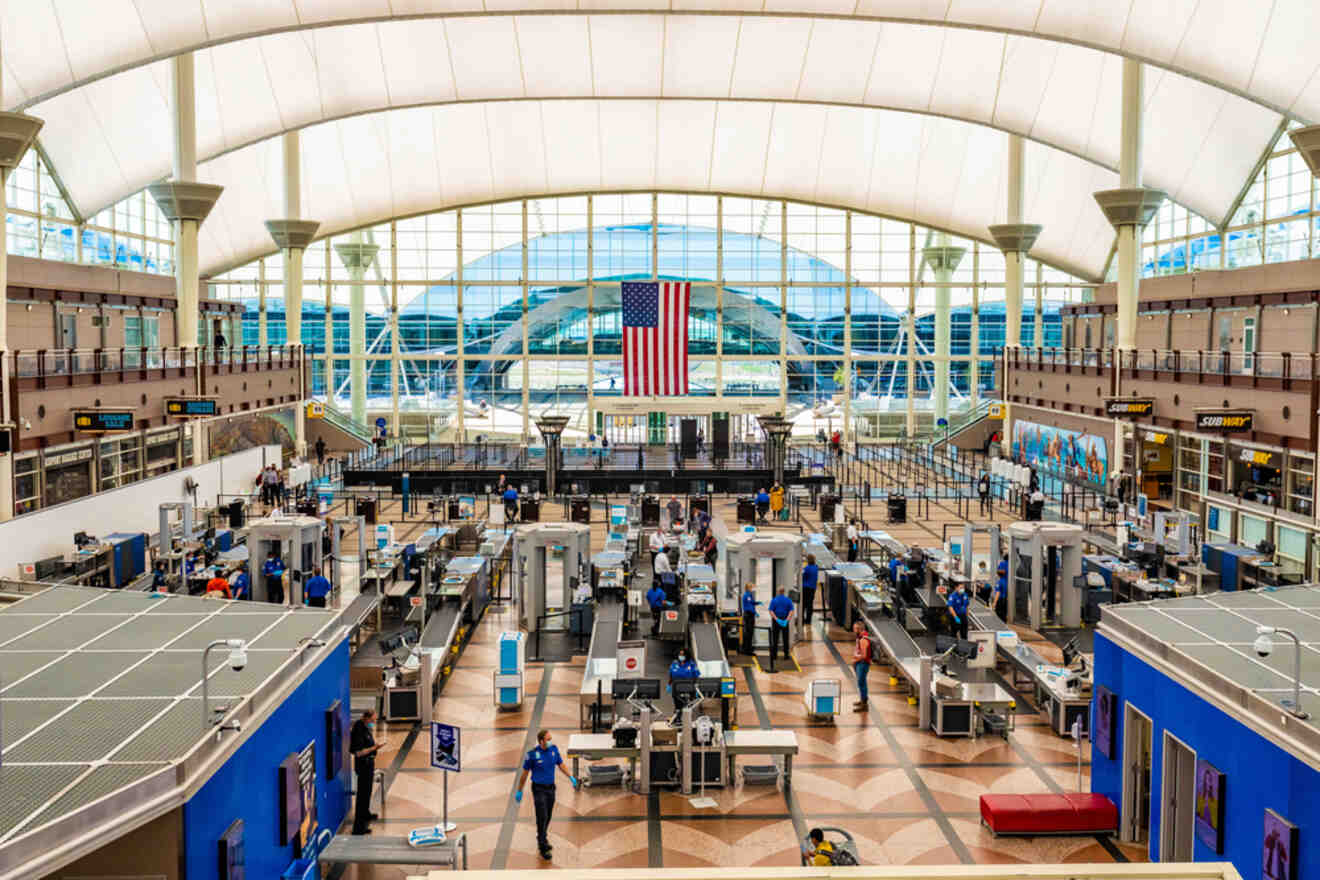 Interior of Denver International Airport with travelers at security checkpoint and a large American flag hanging overhead.