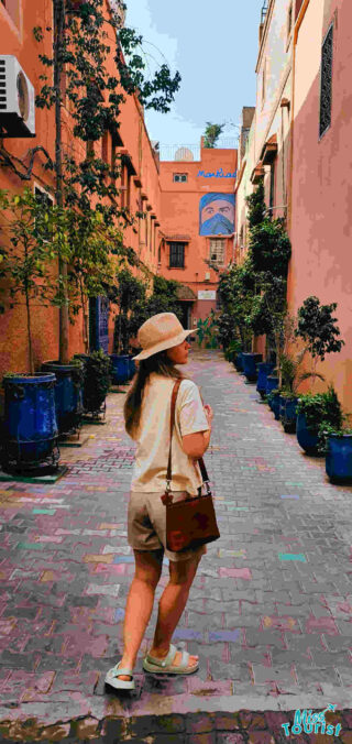 The writer of the post in a sunhat gazes down a colorful alley lined with potted plants and a vibrant mural in Marrakech