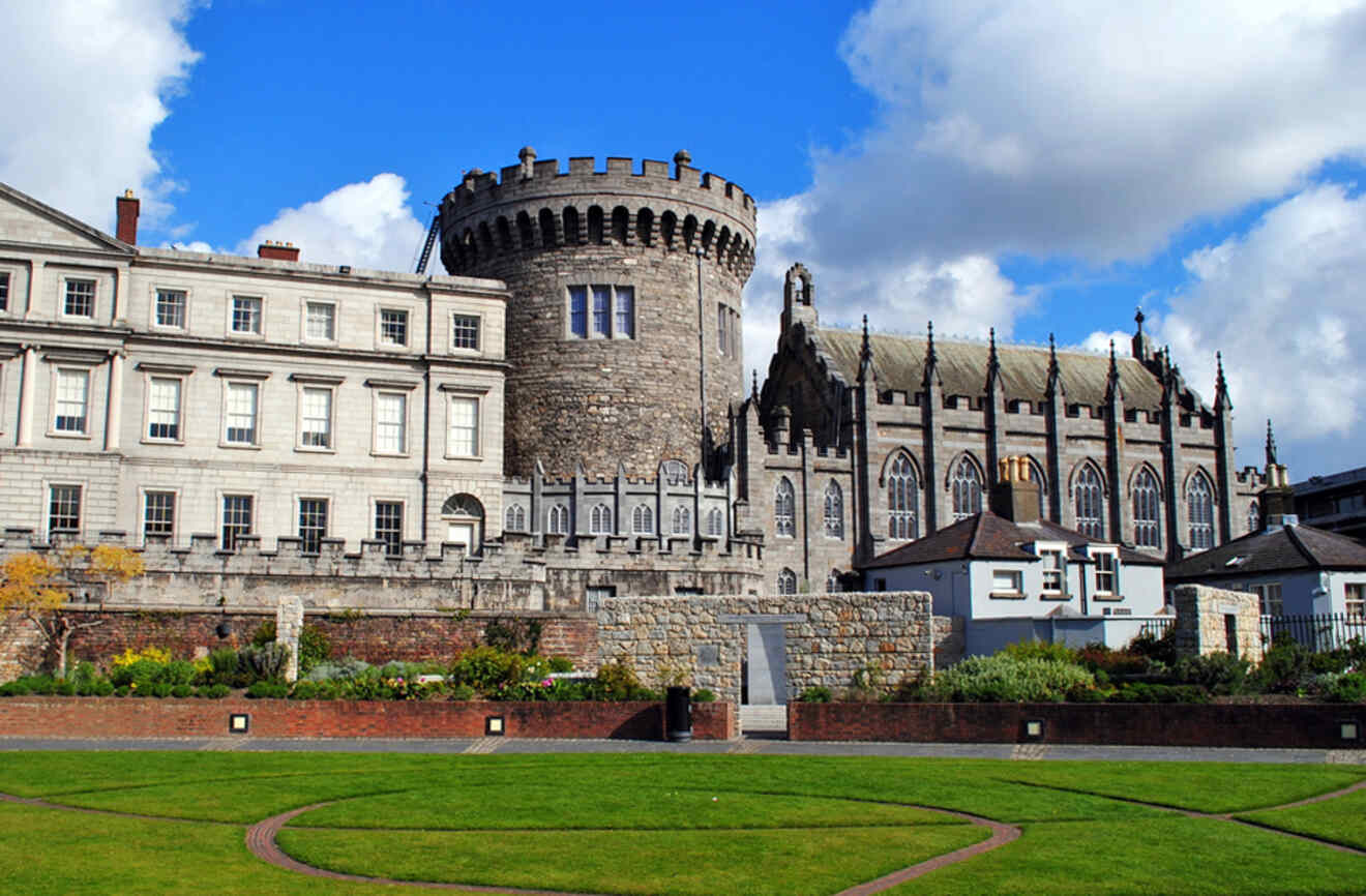Dublin Castle against a blue sky, displaying a mix of medieval and Georgian architecture surrounded by manicured lawns and modern city elements.