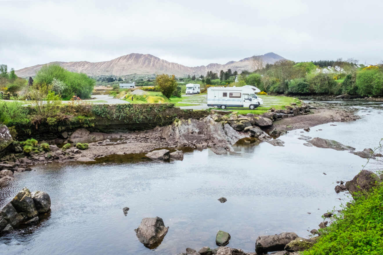 A serene riverside scene with a campervan parked in the distance, surrounded by a lush green landscape and mountains, capturing the peacefulness of campervan travels in Ireland.