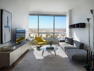 Modern living room with large windows, a gray sofa, yellow armchair, wall-mounted TV, round coffee table, and city view in the background.