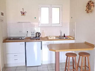 Cozinha pequena com armários brancos, frigobar, pia, fogão, chaleira e dois bancos de madeira em um balcão sob uma janela.