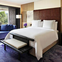 Um quarto de hotel moderno com uma cama impecavelmente arrumada com lençóis brancos, um banco almofadado aos pés da cama, um assento junto à janela e uma grande janela com vista para a vegetação exterior.