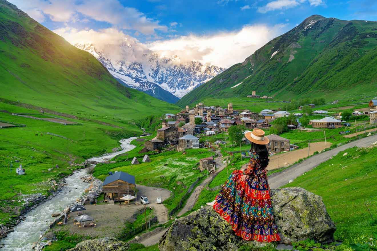 A woman in a vibrant, traditional dress and a wide-brimmed hat stands facing a picturesque mountain village. The lush green landscape, flowing river, and snow-capped peaks in the distance highlight the tranquil beauty of rural life