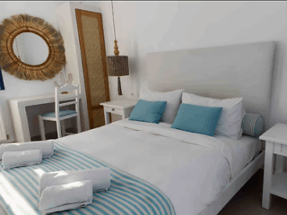 A neatly made bed with blue and white linens, two blue pillows, and three rolled towels; two white nightstands, a vanity with a mirror, and a hanging lamp are in the room.
