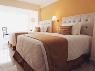 Duas camas impecavelmente arrumadas, com roupa de cama branca e detalhes em marrom, estão dispostas contra uma parede amarela em um quarto de hotel luminoso e moderno, com iluminação contemporânea e uma cadeira junto à janela.