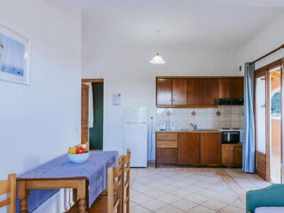 Cozinha e sala de jantar pequenas, com armários de madeira, mesa com cadeiras, fruteira, geladeira e porta aberta para o exterior.