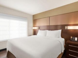 Hotel room with a neatly made bed, two pillows, a wooden headboard, two wall-mounted lamps, a window with blinds, and a nightstand with a clock and phone.