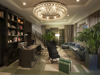 Modern living room with a large chandelier, blue sofa, black chairs, desk, bookshelves, potted plants, stairway, and patterned floor.