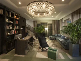 Modern living room with a large chandelier, blue sofa, black chairs, desk, bookshelves, potted plants, stairway, and patterned floor.