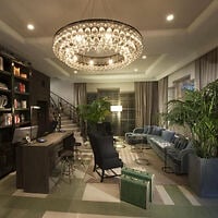 Modern living room with a large chandelier, bookshelves, a desk, blue sofa, armchairs, potted plants, and a suitcase on a patterned rug. Stairs and windows are visible in the background.