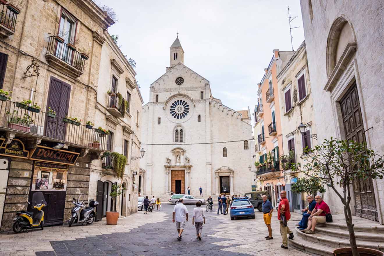 An inviting cobblestone street in Bari, Italy, leading to a majestic cathedral with a rose window, flanked by historic buildings with charming balconies and locals enjoying the day.