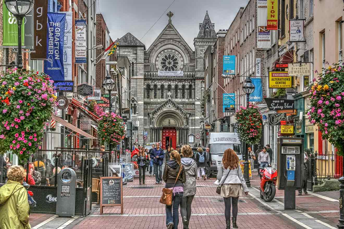 A vibrant street scene in Dublin, with people walking, abundant flower baskets, and a mix of shops leading to an ornate stone church facade.
