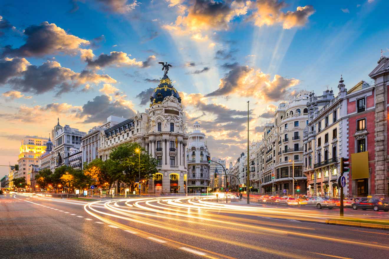 A bustling street in Madrid at dusk, with long exposure capturing streaks of light from moving traffic. Historic buildings with intricate facades line the avenue, while the sky above shows a dynamic pattern of clouds illuminated by crepuscular rays.