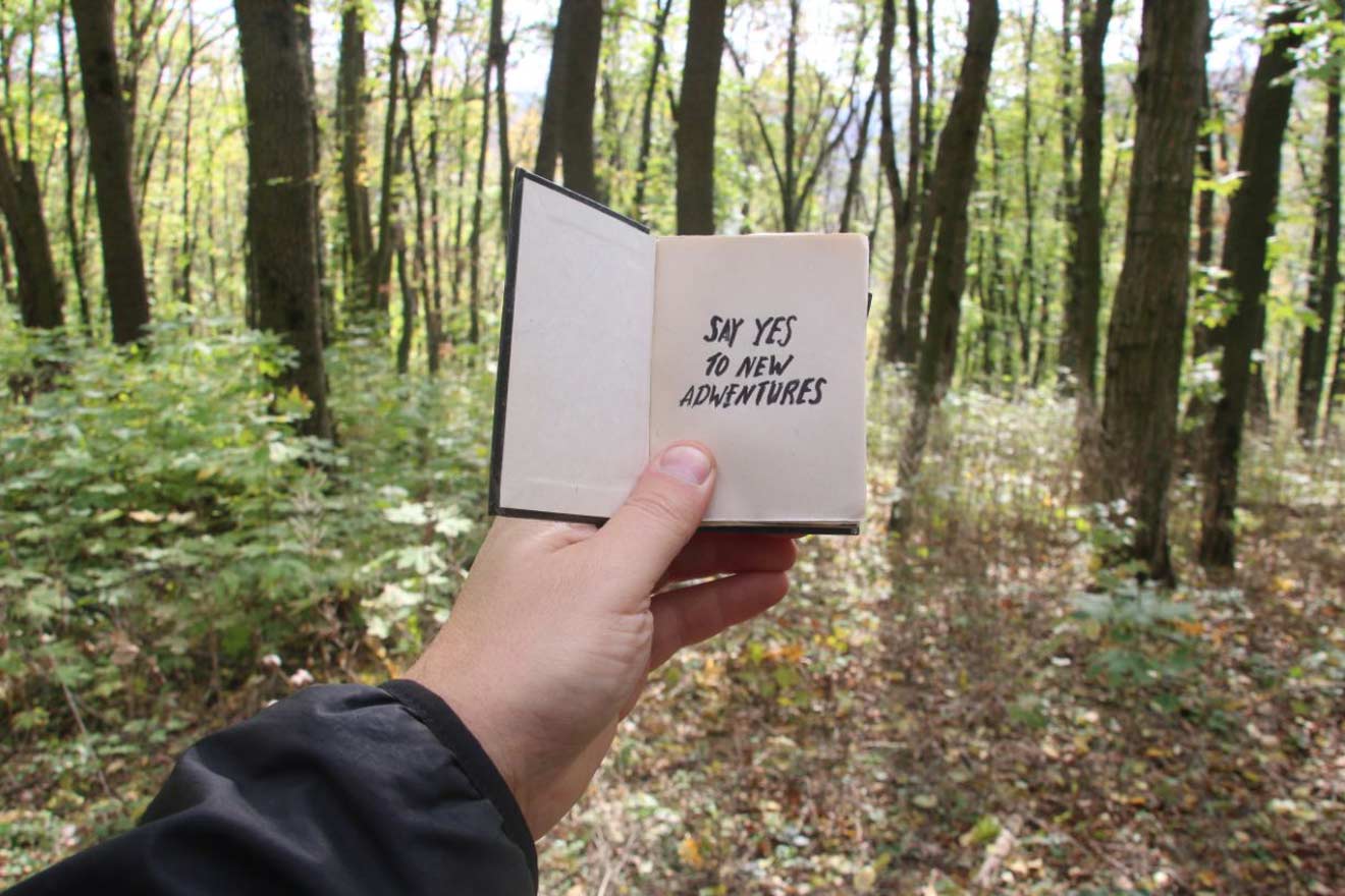 A hand holding an open notebook in a forest, with the phrase 'SAY YES TO NEW ADVENTURES' written on the right page, embodying a spirit of exploration and travel