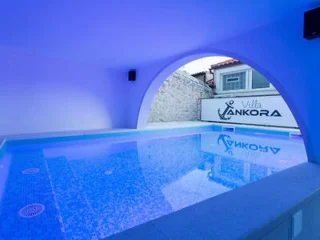 Indoor swimming pool with blue lighting, tiled walls, and an arched opening revealing a sign that reads "Villa Ankora" outside.