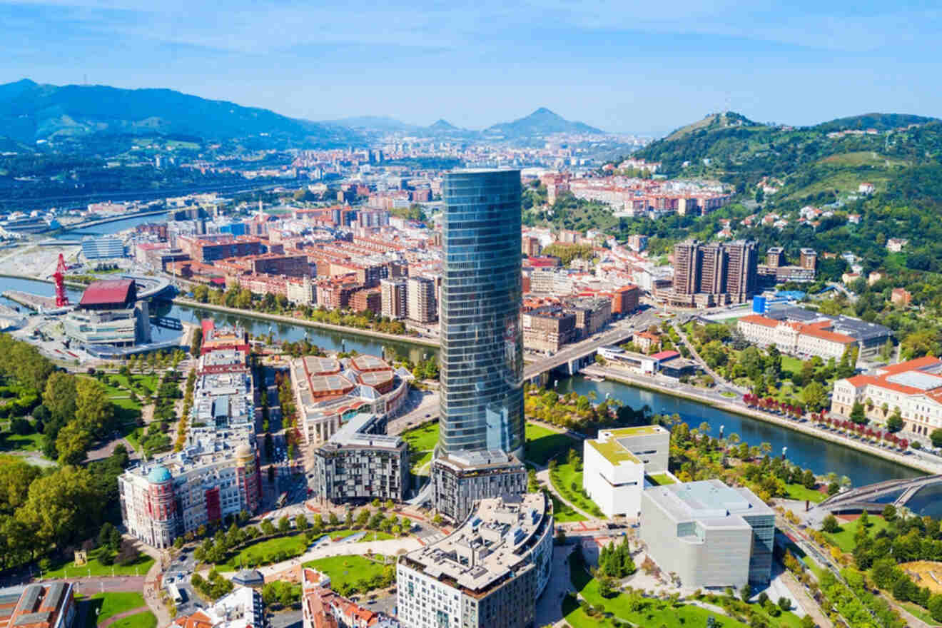 Aerial view of Bilbao with a tall glass skyscraper near a river, surrounded by various buildings and green hills in the background.