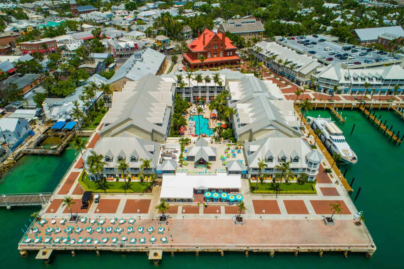 Aerial view of a waterfront resort with a pool surrounded by palm trees, lounge chairs, and cabanas, and a marina with docked boats to the right. There is a red-roofed building in the background and a street with parked cars and colorful buildings on the left.