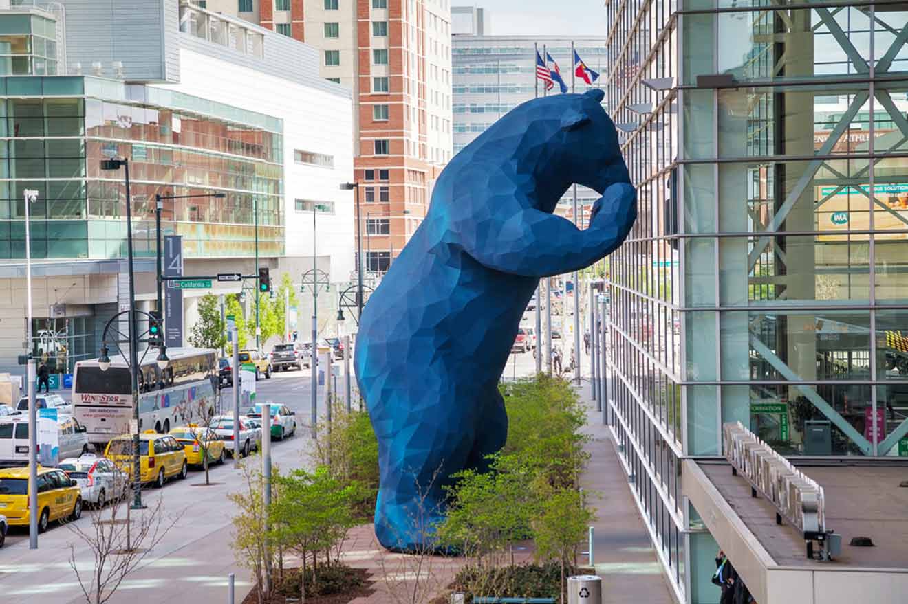 A colossal blue bear sculpture peering curiously into a glass-walled convention center, set against the backdrop of a bustling Denver street with taxis and buses.