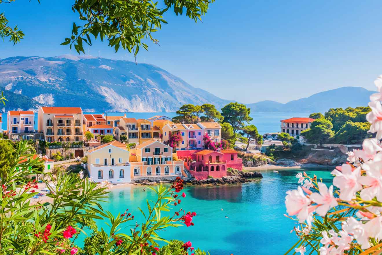 Colorful coastal town with terracotta roofs overlooking a serene blue bay, framed by pink oleander flowers in the foreground and a majestic mountain backdrop