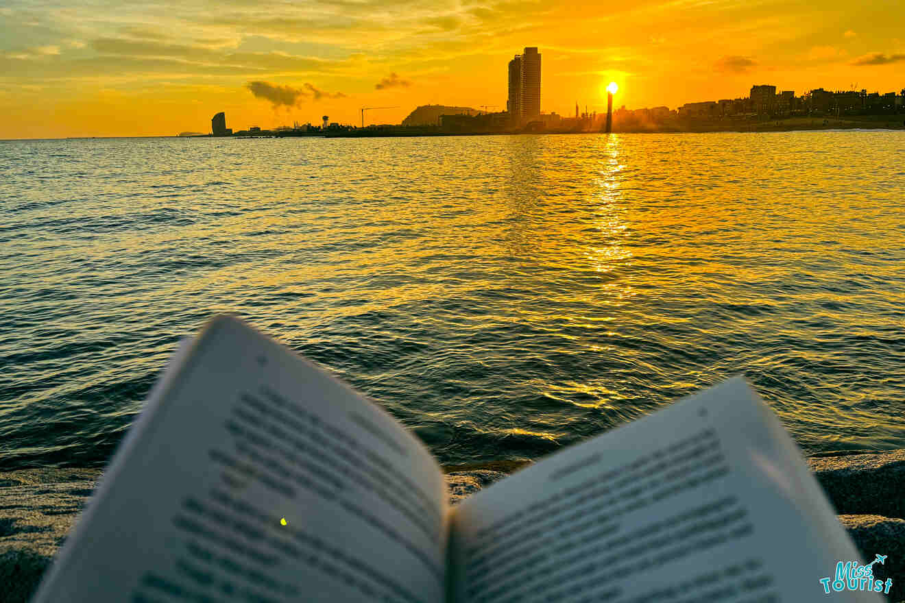 Open book on a sandy beach at sunset with the sun reflecting on the water's surface, and the silhouette of a Barcelona skyline in the background