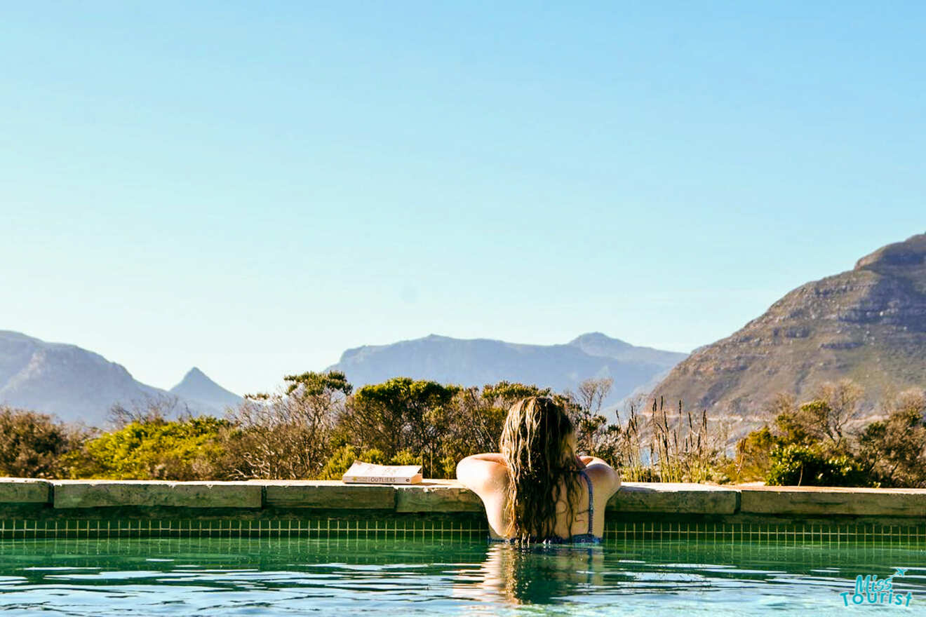 Author of the post with long hair sitting at the edge of an outdoor infinity pool, looking out towards a mountainous landscape under a clear blue sky