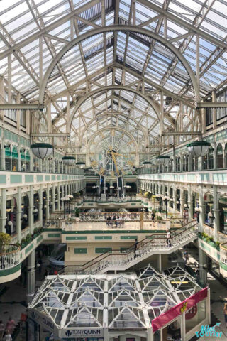 An elegant interior of the St. Stephen's Green Shopping Center, showcasing a glass roof, multiple shopping levels with ornate railings, and a central clock feature.