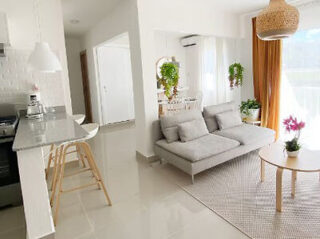 Sala de estar moderna e iluminada com sofá cinza, mesa de centro redonda, balcão de cozinha com banquetas, plantas suspensas e em vasos, e grandes janelas com cortinas laranja.